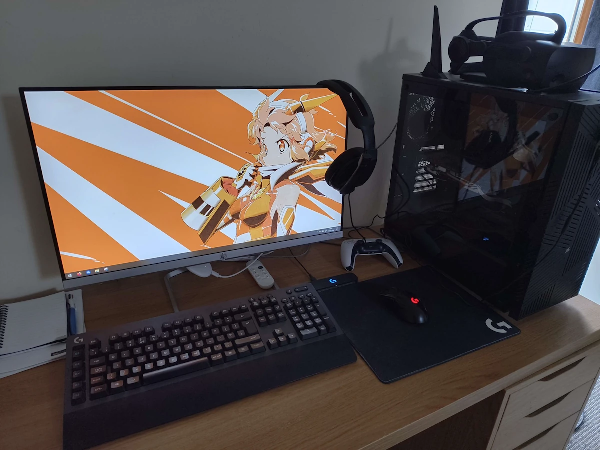 A photograph of a desk. Atop the desk sits a black PC with a glass side, a mouse and keyboard, and a monitor. The monitor is currently idle on the windows desktop, displaying a wallpaper featuring the cover art of the Symphogear XV Hibiki Tachibana album.